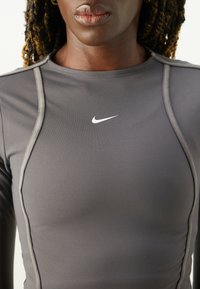 Graues Sportoberteil aus dehnbarem Material, mit figurnahem Design, kontrastierenden Nähten und einem weißen Nike-Logo auf der Brust.