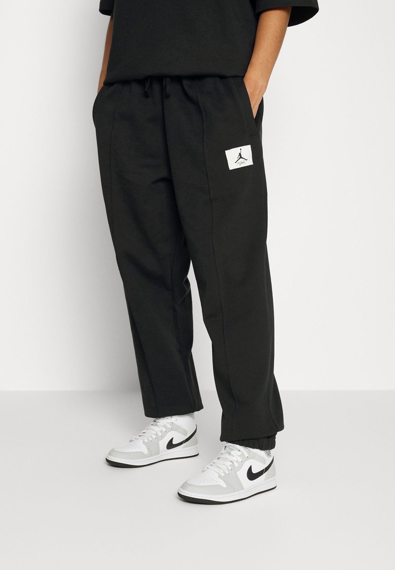 Schwarze Sweatpants mit lockerer Passform, ausgestattet mit einem elastischen Bund und einem weißen Jordan-Logo-Patch am linken Oberschenkel. Kombiniert mit weißen und grauen Nike-Sneakern.