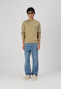 Beige katoenen sweatshirt met het "Club1990" logo, gecombineerd met lichtblauwe jeans en crèmekleurige sneakers. Model draagt donkere zonnebril.