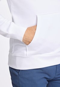 Sudadera blanca de algodón con un bolsillo frontal y puños acanalados, combinada con pantalones azules texturizados que presentan un acabado suave.