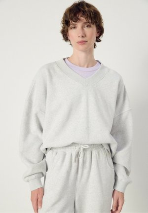 Person with short brown hair wearing light grey V-neck sweatshirt and matching drawstring pants against plain white background.