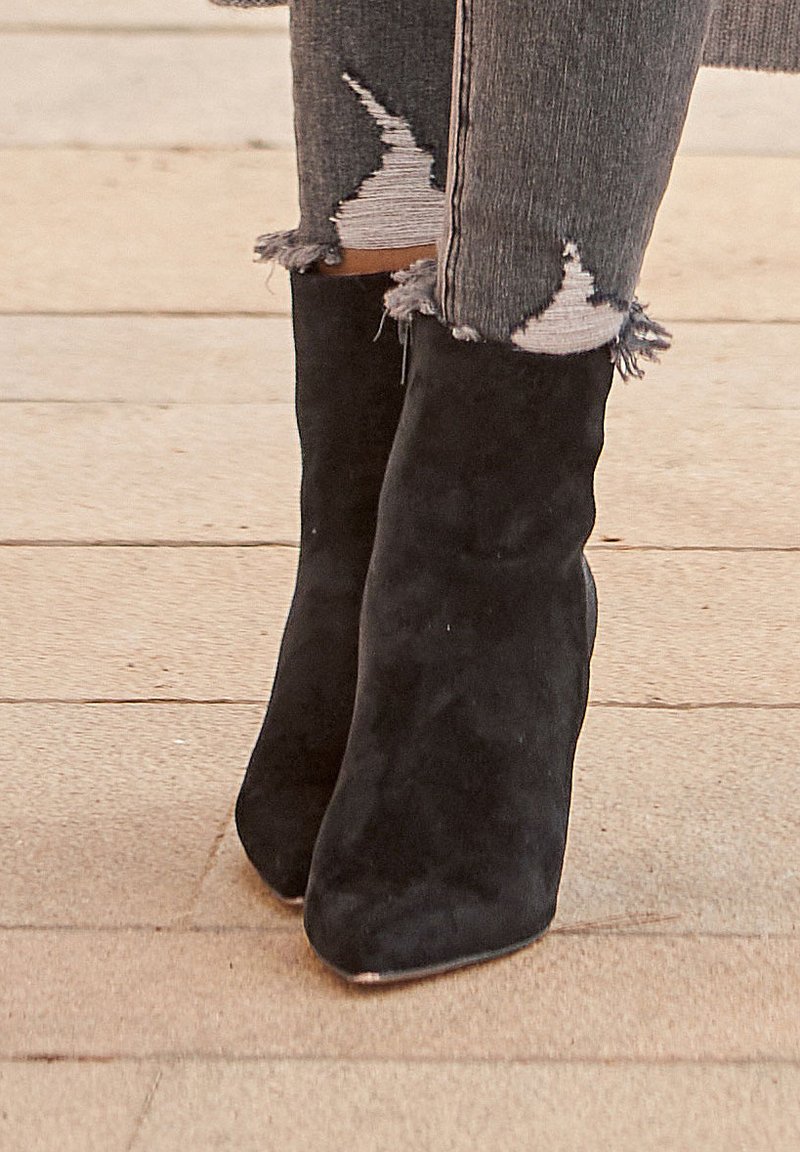 Stivaletti alla caviglia neri in suede con punta affusolata, caratterizzati da una texture liscia e hardware minimale, accanto a jeans grigi usurati con orli sfrangiati.