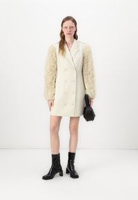 Manteau crème à double boutonnage avec des manches en fourrure texturée, associé à un sac à main géométrique noir et des bottes noires à talons hauts.