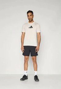 Kerge beež lühikeste varrukatega T-särk musta Adidas logo ja tumedate lühikesed püksid ning mustade spordijalanõudega.