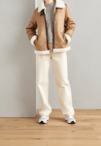Giacca in ecopelle scamosciata marrone con colletto e polsini in shearling bianco, abbinata a pantaloni a gamba larga color crema e sneakers grigie.