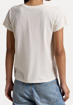 Person iført en ensfarvet hvid kortærmet t-shirt og lyseblå denim-jeans, set bagfra mod en neutral baggrund.