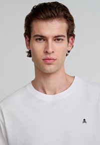 Witte katoenen t-shirt met een ronde halslijn, voorzien van een klein geborduurd doodskop en gekruiste botten logo op de linkerborst.