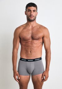 Uomo a torso nudo con capelli corti e scuri, che indossa boxeur grigi con elastico nero etichettato "HUGO", in piedi davanti a uno sfondo bianco semplice.