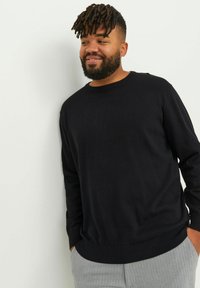 Pulover negru cu mâneci lungi, guler rotund și manșete cu model cu rigle, asortat cu pantaloni gri cu dungi. Materialul pare neted și ajustat.