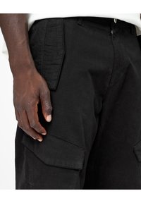 Mano de piel oscura descansando en el bolsillo de pantalones cargo negros con solapa, vistiendo una camisa blanca metida por dentro, primer plano de la cintura y el muslo.