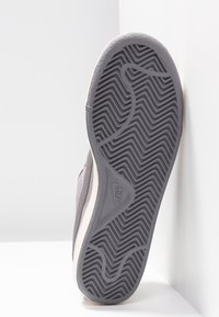 Semelle grise d'une basket Nike avec motif de bande de roulement en zigzag, positionnée debout contre un mur et un sol blancs.