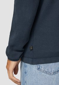 Granatowy, fakturowany sweter z ribbingu na końcach, z małą czarną metką logo z boku, zestawiony z jasnoniebieskimi dżinsami.