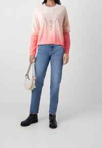 Pull dégradé de rose clair à corail avec un logo triangulaire "GUESS", associé à un jean en denim bleu et des bottines noires; elle tient un sac à main crème.