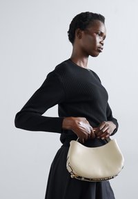 Sac en cuir couleur crème avec une forme arrondie, accentué de garnitures en or, et un pull noir à côtes texturé au premier plan.