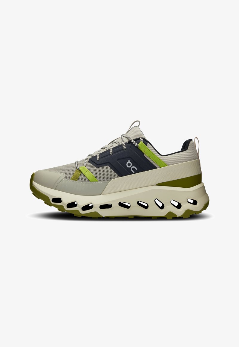 Zapatilla deportiva ligera en beige con acentos en verde y gris oscuro. Parte superior de malla, cuello acolchado, suela gruesa con recortes ovalados.