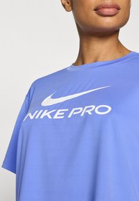Αθλητική μπλούζα Nike Pro σε ανοιχτό μπλε χρώμα, κατασκευασμένη από απαλό ύφασμα, με μεγάλο λευκό λογότυπο και κείμενο στο στήθος, με κοντά μανίκια.