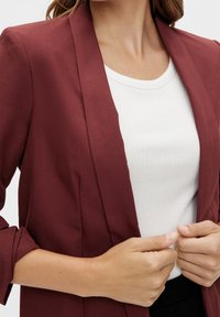 Blazer bordeaux à coupe ajustée, col à revers crantés et manches retroussées, porté sur un haut blanc côtelé. Texture du tissu lisse.