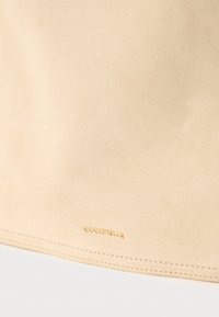 Επιφάνεια μπεζ σουέντ με λεπτή ραφή κατά μήκος της άκρης και μικρό χρυσό λογότυπο «COCCINELLE» ανάγλυφο κοντά στο κάτω κέντρο.