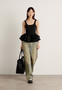 Čierny top bez rukávov s vrstveným volánom na okrajoch, v kombinácii so svetlozelenými džínami. Modelka nesie veľkú čiernu tote tašku. Jednoduché pozadie.