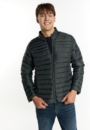 Ung mand smilende iført en mørkegrøn quiltet jakke og blå jeans, stående med hænderne nær lommerne mod hvid baggrund.