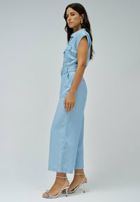 Combinaison en denim bleu clair avec des manches courtes, fermeture à boutons sur le devant, taille ceinturée et jambes larges. Associée à des talons argentés à lanières.