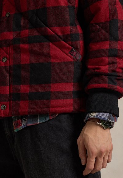 Sarkani un melna pārbaudes flaneļa jaka ar šūtu faktūru, melnām ribainām aprocēm un melnu plāksteri ar logotipu. Valkāta pār daudzkrāsainu pārbaudes kreklu.