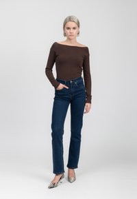 Braunes, schulterfreies, figurbetontes Oberteil, blaue, hoch taillierte Jeans und spitze, mit Schlangenledermuster verzierte Absätze, vor einem neutralen Hintergrund.