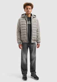 Lichtgrijze gewatteerde jas met horizontale stiksels, ritssluiting aan de voorkant en capuchon, gedragen over een donkere trui en zwarte jeans, met sneakers.