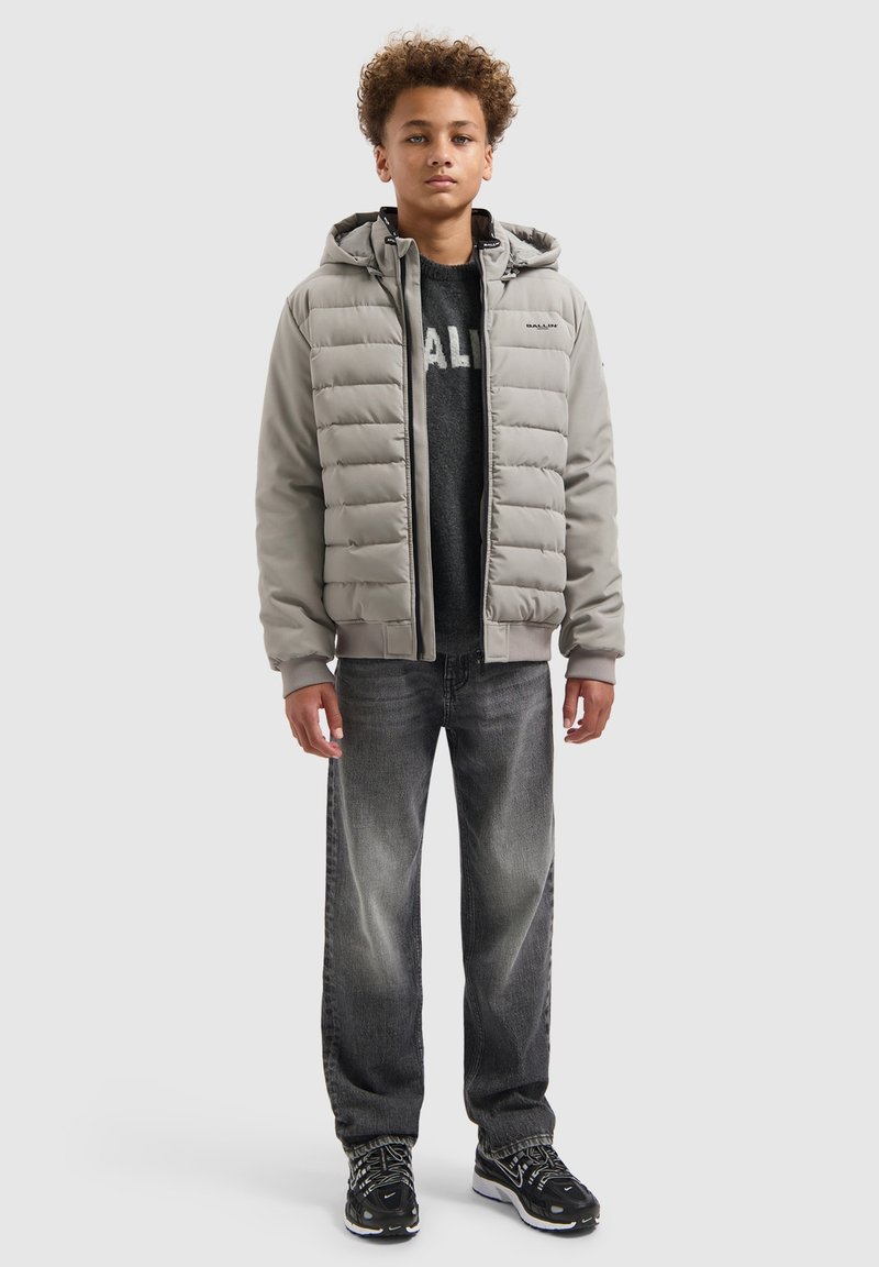 Lichtgrijze gewatteerde jas met horizontale stiksels, ritssluiting aan de voorkant en capuchon, gedragen over een donkere trui en zwarte jeans, met sneakers.