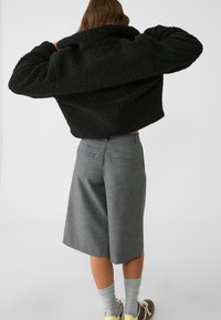 Musta lyhyt pörröinen takki, jossa on ylisuurikokoinen kaulus, yhdistetty harmaisiin mittatilaustyönä tehtyihin shortseihin ja nilkkapitkiin sukkiin. Kankaan pinta on teksturoitu, ja takissa on käytännölliset taskut.