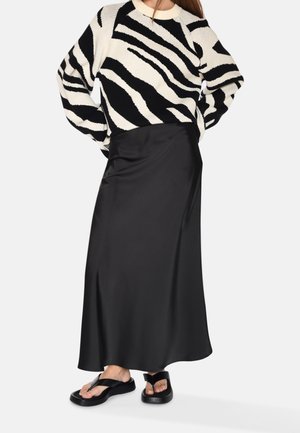 Femme porte un pull rayé zèbre noir et blanc, une longue jupe en satin noire et des sandales compensées noires avec lanières entre les orteils.