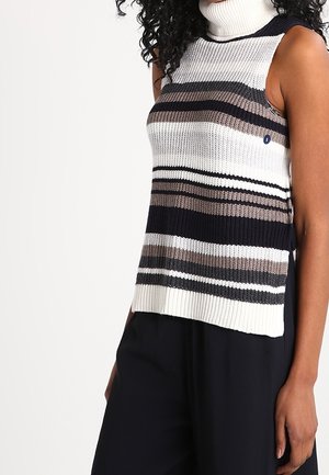 Frau trägt einen ärmellosen, gestreiften Strick-Rollkragenpullover in Schwarz-, Weiß- und Brauntönen, kombiniert mit schwarzen Hosen.