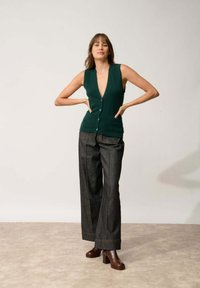 Zielony, bezrękawnikowy sweter z dekoltem w kształcie litery V i zapięciem na guziki, w połączeniu z szerokimi ciemnoniebieskimi spodniami dżinsowymi i brązowymi botkami na obcasie.