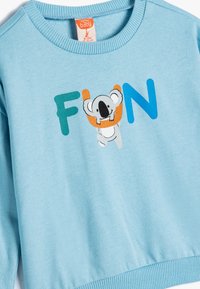Sudadera de color azul claro con cuello y dobladillo acanalados, que presenta la palabra "DIVERTIDO" en letras coloridas y un koala de dibujos animados colgando de la letra "U".