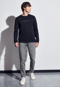 Man in een zwart sweatshirt met de tekst "Crafted Goods", grijze jeans en witte sneakers, staand binnen tegen een witte muur.