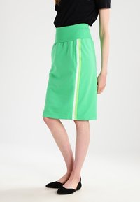 Green athletic skirt with a wide, ribbed waistband. Features a side stripe in light green and white. Fabric appears smooth and stretchy.