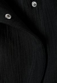 Gros plan sur un tissu noir texturé avec un bouton pression argenté visible. Surface douce et légèrement froissée avec des détails de couture prononcés.