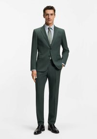 Dunkelgrüner Anzug aus strukturiertem Stoff, schmal geschnitten, kombiniert mit einem hellblauen Hemd und einer gemusterten Krawatte. Schwarze Anzugschuhe.