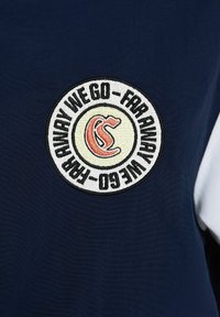 T-shirt bleu marine avec un écusson circulaire brodé comportant du texte et un logo en rouge et jaune, sur un fond de tissu lisse.