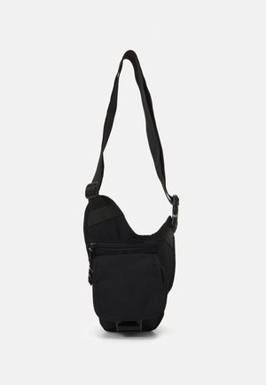 Petit sac noir porté en bandoulière avec sangle réglable, poche avant zippée et fermeture à rabat, présenté suspendu sur un fond uni.