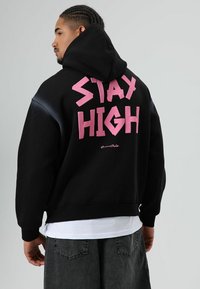 Zwarte hooded sweatshirt met grote roze tekst 'STAY HIGH' op de achterkant. Voorzien van een comfortabele pasvorm en geribbelde boorden. Draagt over een wit T-shirt.