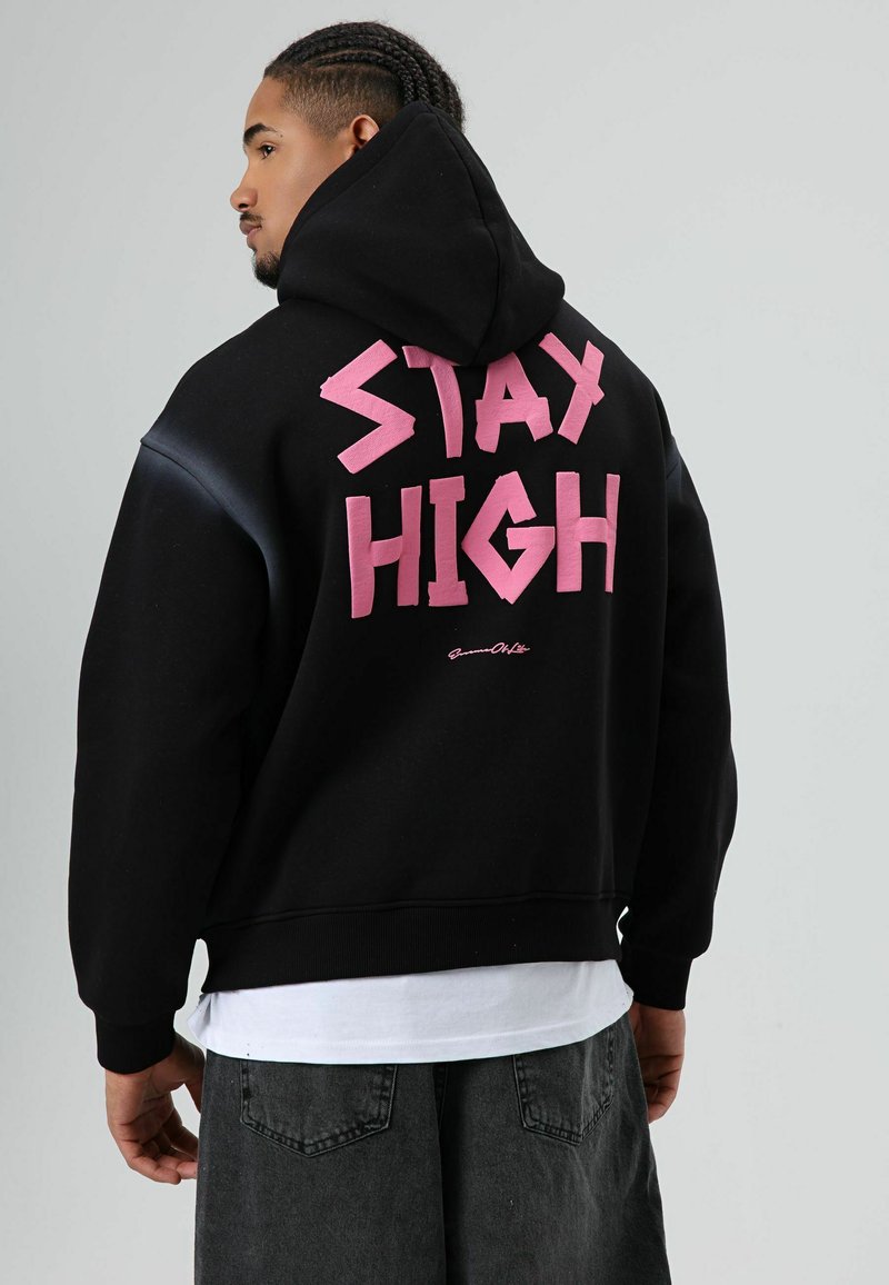Zwarte hooded sweatshirt met grote roze tekst 'STAY HIGH' op de achterkant. Voorzien van een comfortabele pasvorm en geribbelde boorden. Draagt over een wit T-shirt.