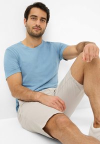 Lichtblauwe katoenen t-shirt, korte mouwen, ronde hals; gecombineerd met beige shorts. Model zit in een casual pose tegen een witte achtergrond.