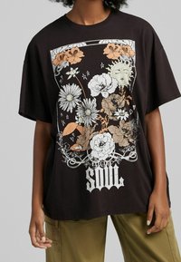 Musta ylipitkä t-paita, jossa on valkoinen, oranssi ja vihreä kukka- ja aurinkokuvio, sekä "ELECTRIC SOUL" -teksti alareunassa.