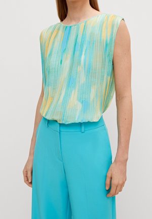 Woman wearing sleeveless pleated top with blue, green, and yellow abstract pattern tucked into high-waisted bright turquoise pants.