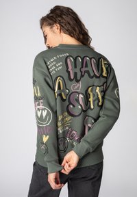 Olive green sweatshirt featuring playful graphic text and patterns in pink, yellow, and black. Ribbed cuffs and hem. Soft fabric texture.