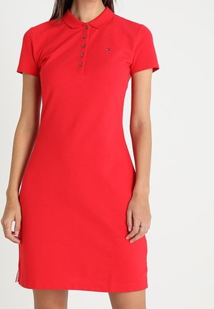 Frau trägt ein eng anliegendes, kurzärmeliges rotes Polo-Kleid mit geknöpftem Kragen und kleinem Logo auf der Brust, steht vor weißem Hintergrund.