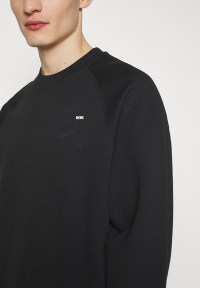 Sweatshirt noir à col rond, coutures d'épaules discrètes et petit logo blanc sur la poitrine, fabriqué en tissu doux et texturé.