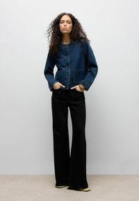 Donkerblauwe denimjack met een ronde halslijn, knoopsluiting en twee zakken. Zwarte wijde broek en crèmekleurige schoenen.