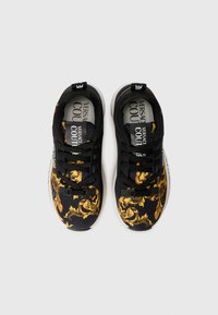 Baskets noires avec un motif orné jaune, dotées de lacets noirs et d'un matériel ajustable. Semelles blanches avec du texte imprimé à l'intérieur.